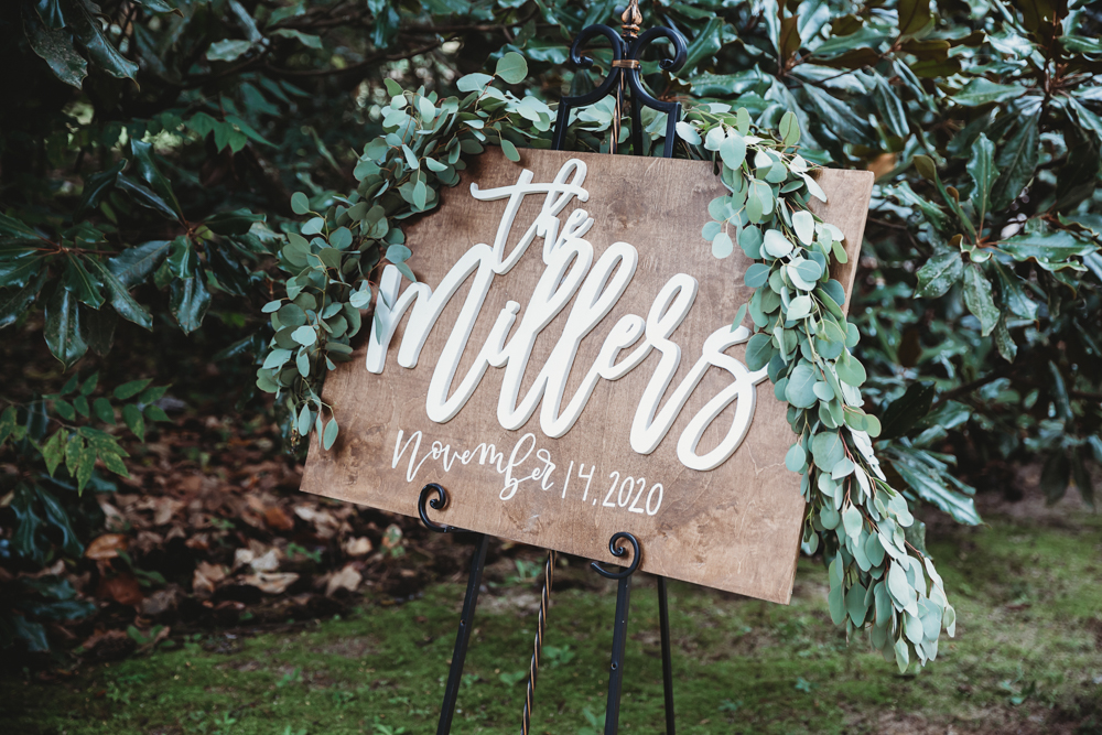 Wooden welcome sign with 'The Millers' and wedding date, accented by eucalyptus garland and forest backdrop.