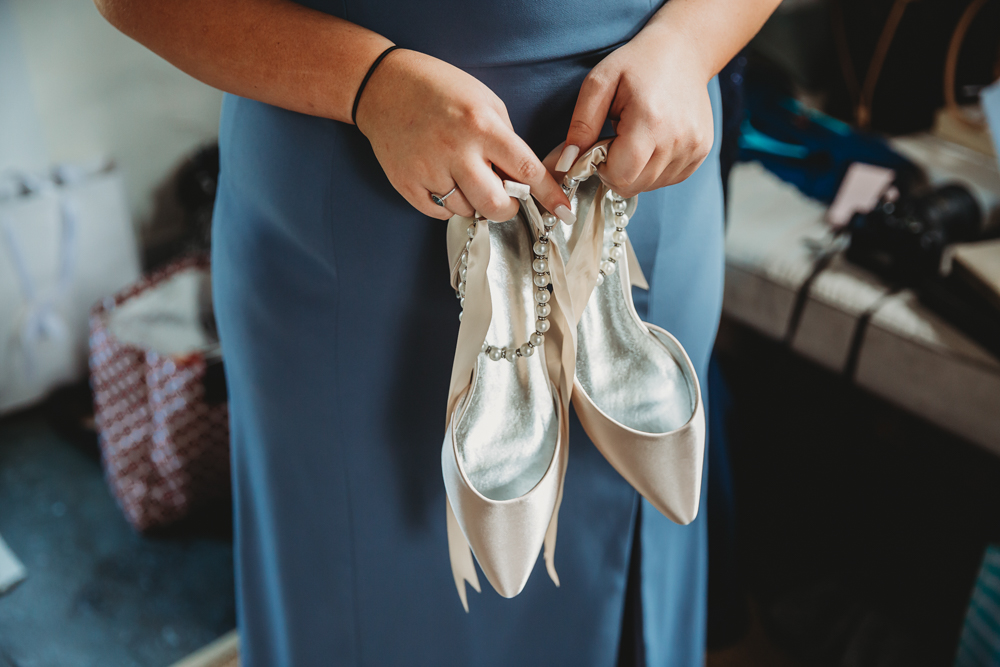 Bridesmaid in slate blue dress holding satin heels with ankle pearls inside a getting-ready suite.
