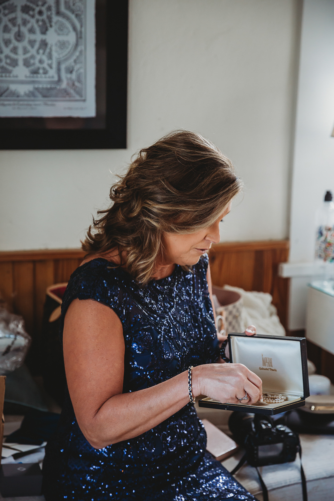 Mother of the bride in navy sequin dress opening a pearl necklace box in the prep room before the ceremony.