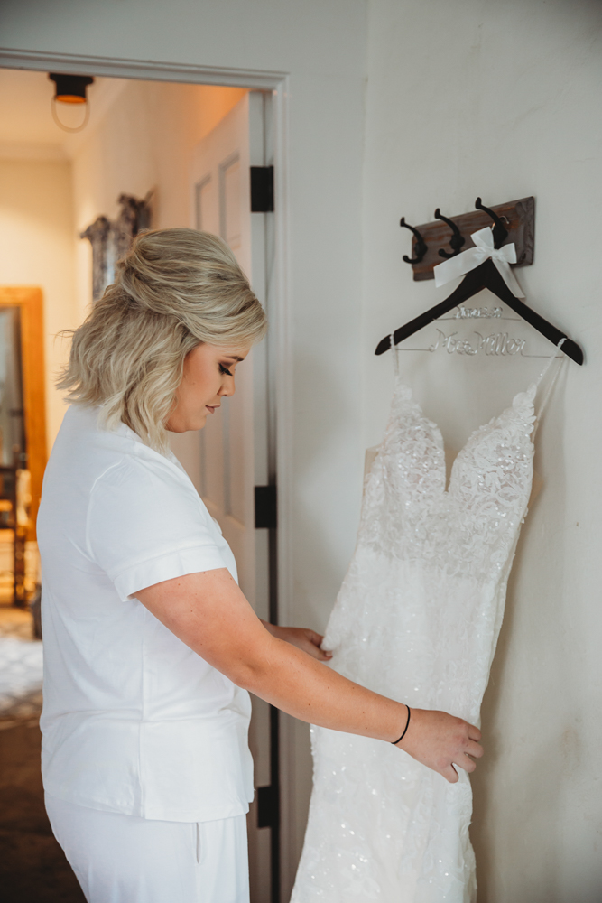 Bride in white pajamas holding lace wedding gown on personalized hanger in softly lit prep room.