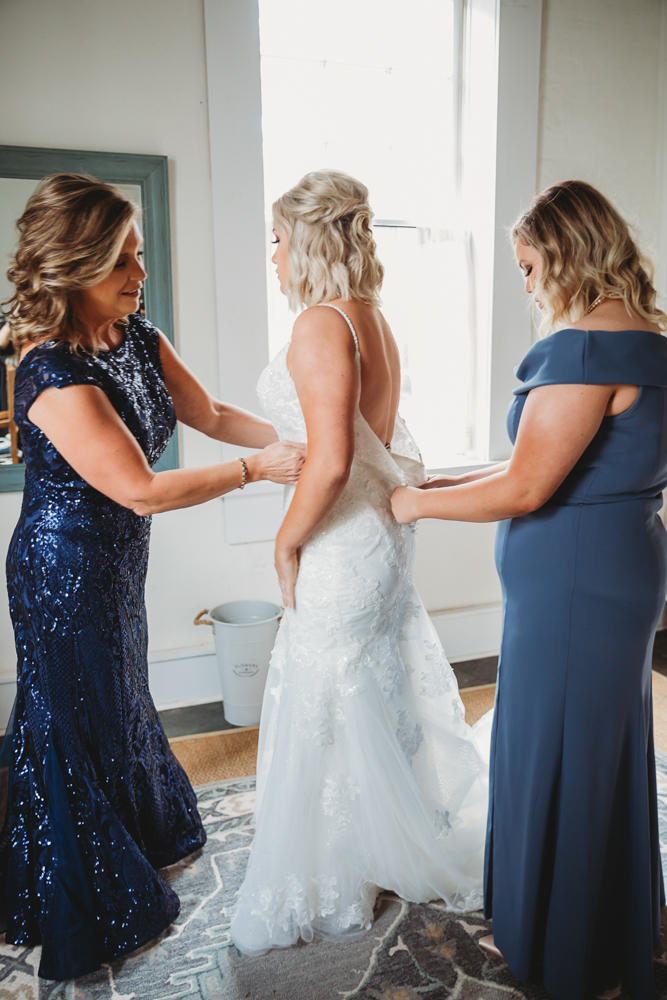 Bride being zipped into gown by mother and bridesmaid, with window light and patterned rug in prep suite.