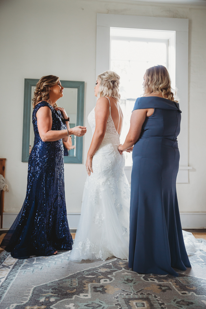 Bride standing while mother and bridesmaid adjust gown details under soft light from tall windows.