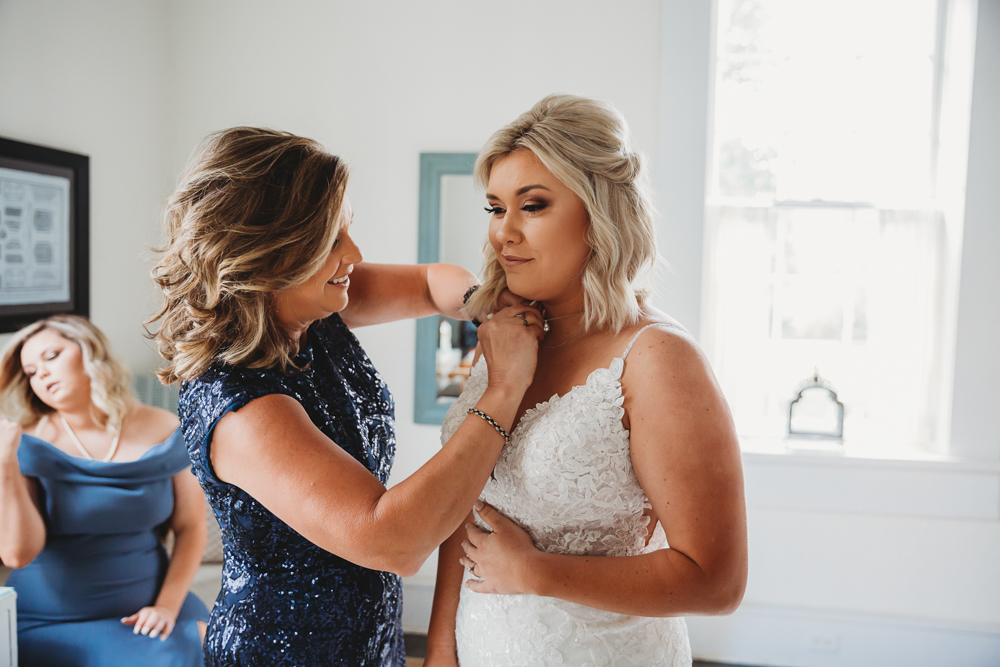 Mother of the bride fastening necklace on her daughter in white lace gown inside a sunlit dressing room.