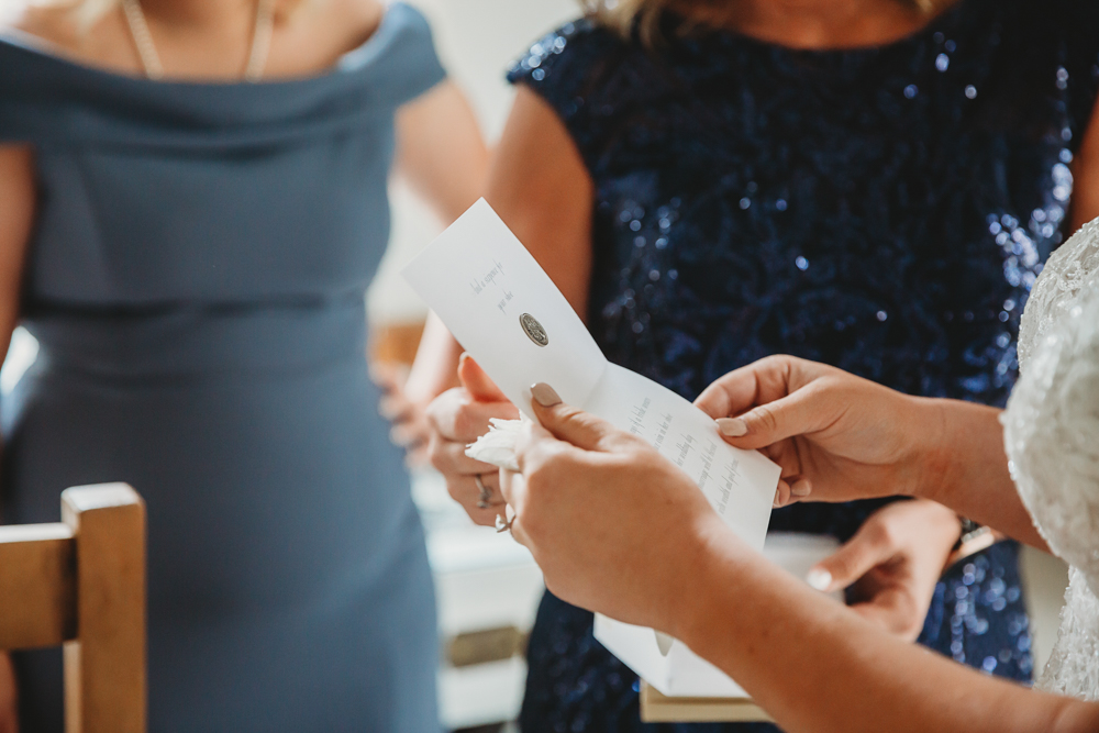 Bride reading a heartfelt note, flanked by women in navy and slate blue dresses, hands and paper in focus.