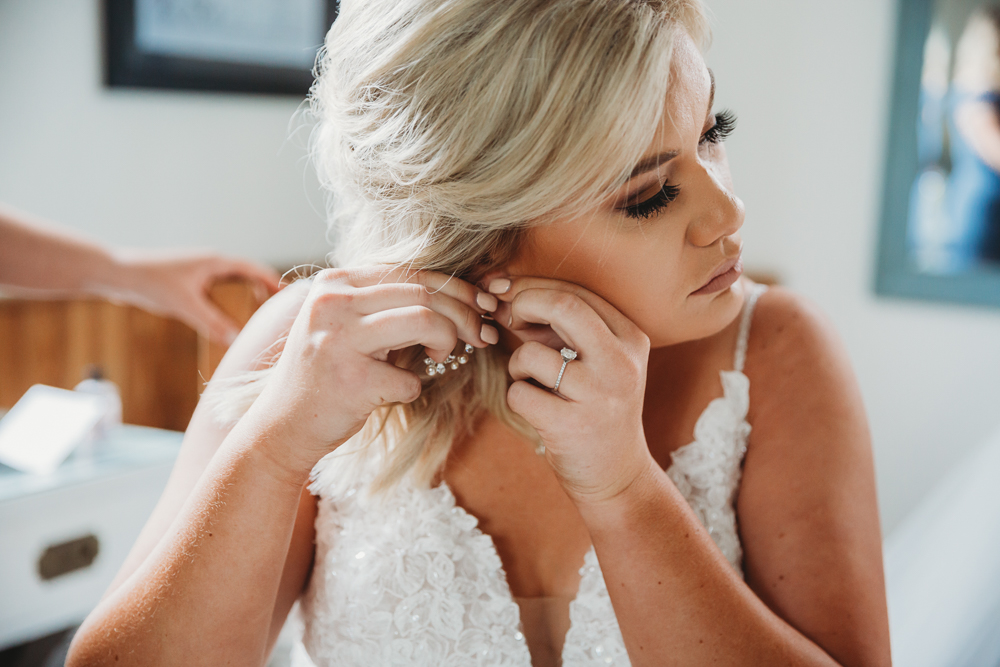 Bride putting on earrings in white lace gown, showing soft curls, bridal glam, and solitaire engagement ring.