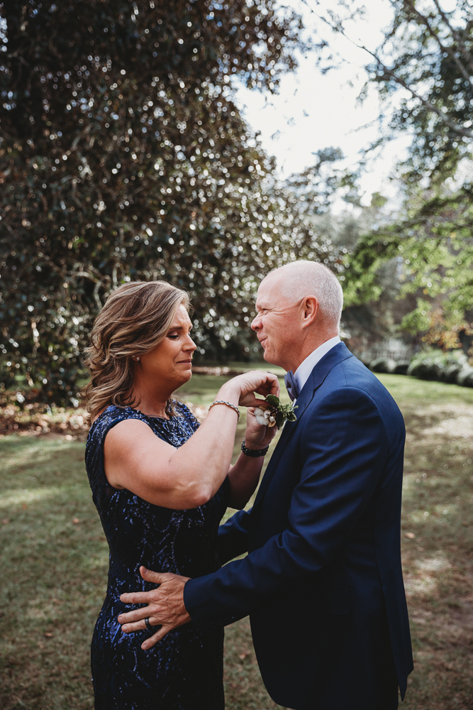 Mother pinning boutonniere on father outside in garden, both smiling in formal navy attire under trees.