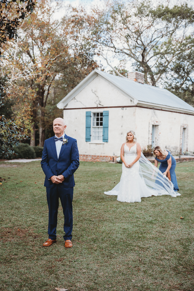 Father of the bride in blue suit standing with hands clasped as bride approaches during emotional first look.