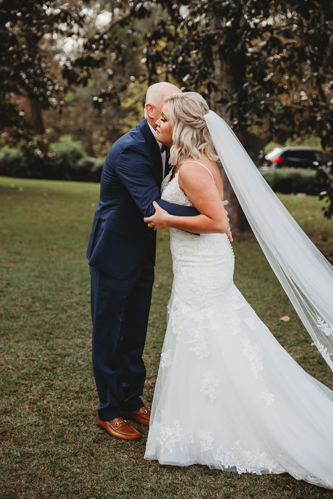 Bride and father embrace during first look outdoors, long veil trailing behind lace gown under trees.