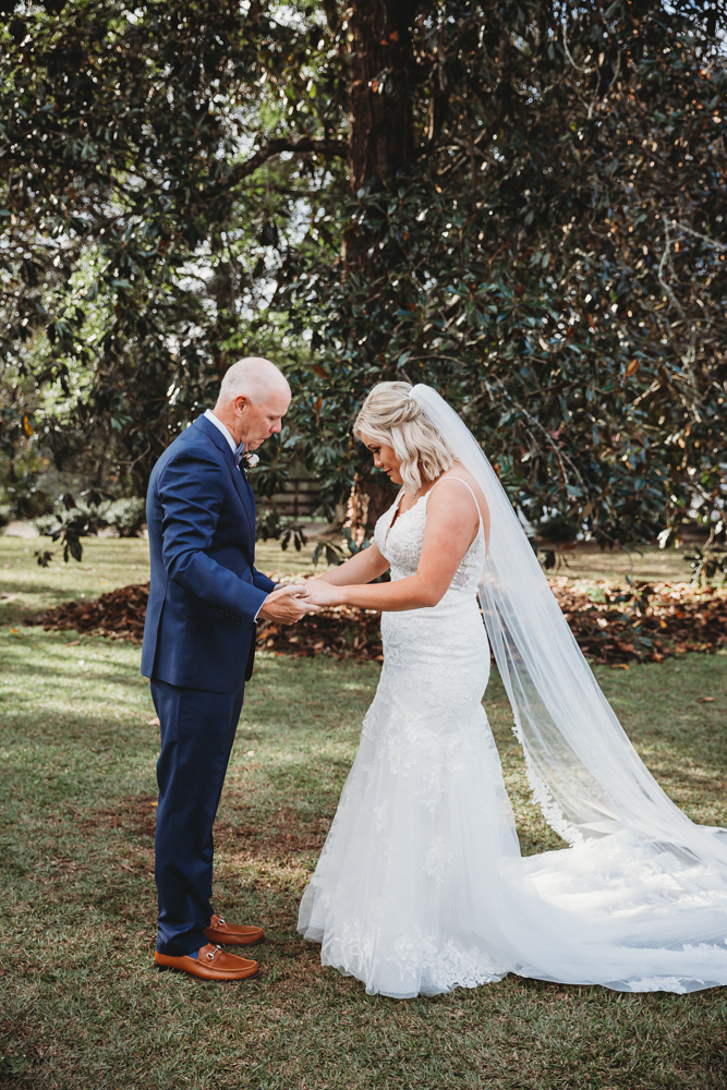 Bride and father holding hands during emotional first look under the tall trees at Wavering Place.