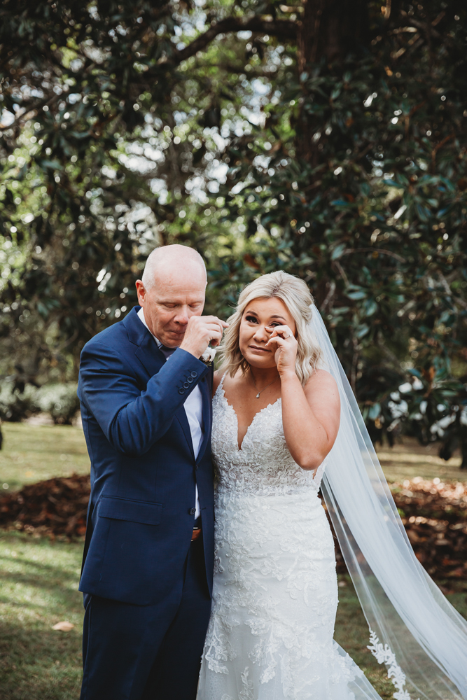 Bride and her father wipe tears side by side in heartfelt outdoor moment beneath the oaks.