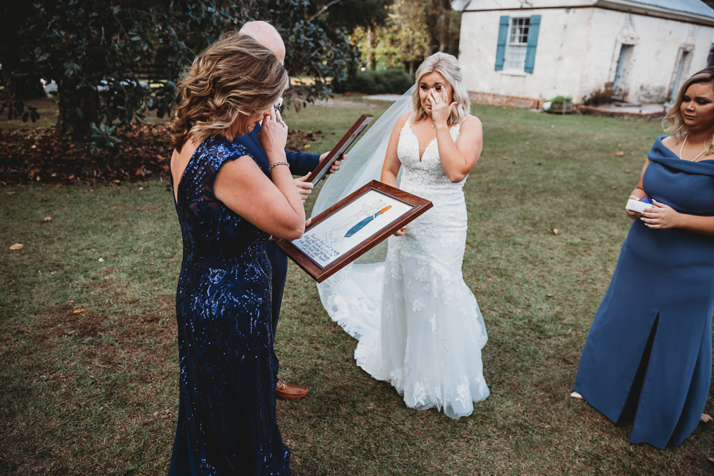 Bride reacting tearfully to a framed wedding illustration presented by her family outdoors.