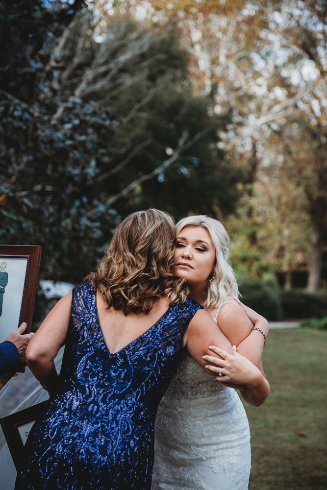 Bride in fitted lace gown hugs her mother in navy sequin gown after special family moment.