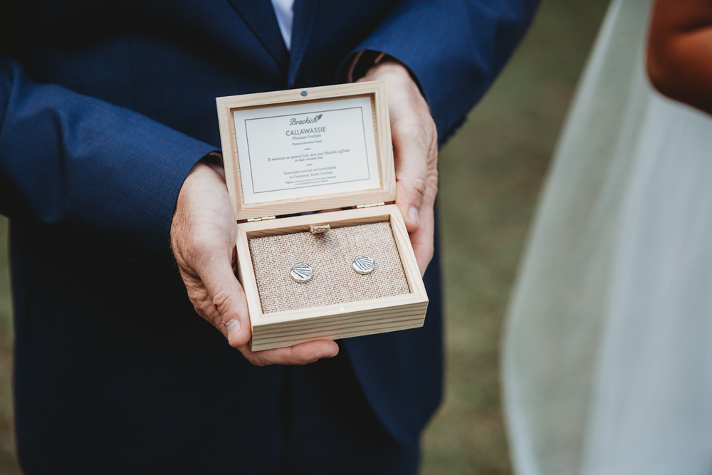 Close-up of wooden box holding monogrammed cufflinks presented in father of the bride’s hand, suit jacket visible.