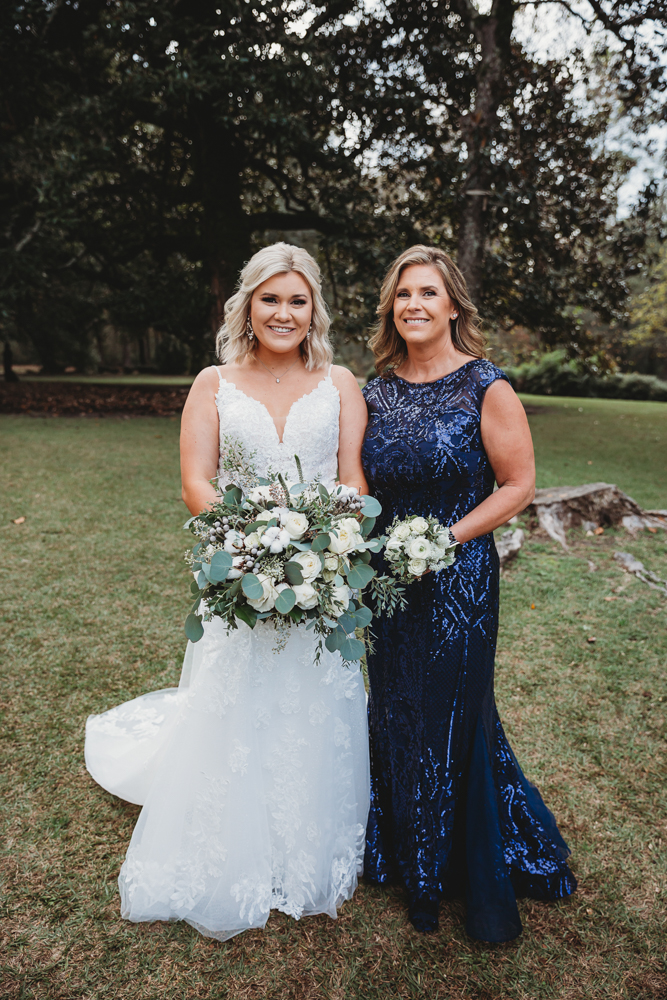 Bride and mother stand smiling side by side with ivory floral bouquets and trees behind them.
