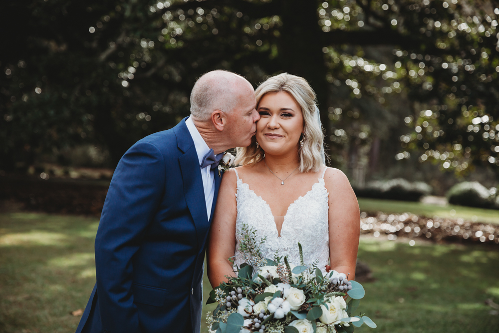 Bride holding bouquet as her father kisses her forehead in a tender moment with greenery behind.
