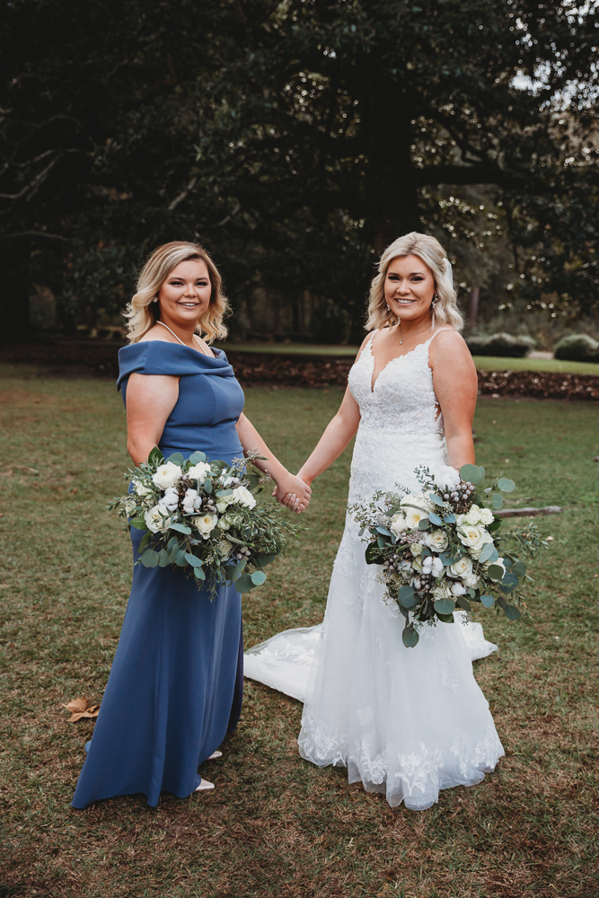 Bride and bridesmaid holding hands in garden wearing white and slate blue gowns with matching bouquets.