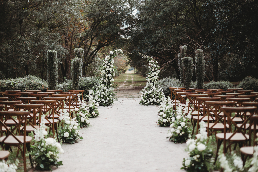 Outdoor ceremony setup with aisle lined in white florals, wooden chairs, and floral arch under trees.