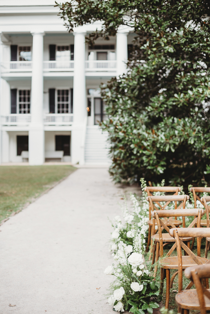 Wooden cross-back chairs with white floral aisle arrangements facing the grand white-columned house at Wavering Place in Eastover SC.
