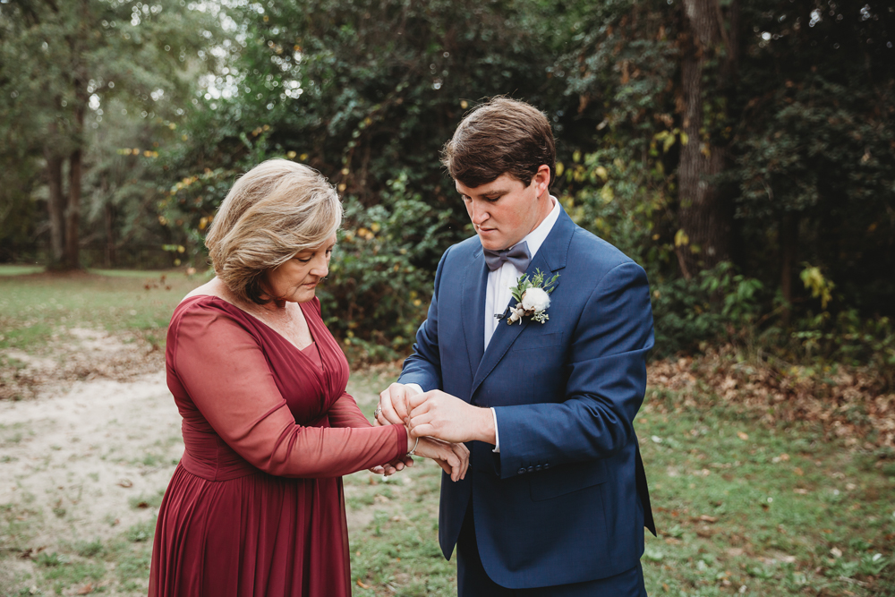 The groom helps his mom with her bracelet outdoors, both dressed formally with trees in the background at Wavering Place in Eastover SC.
