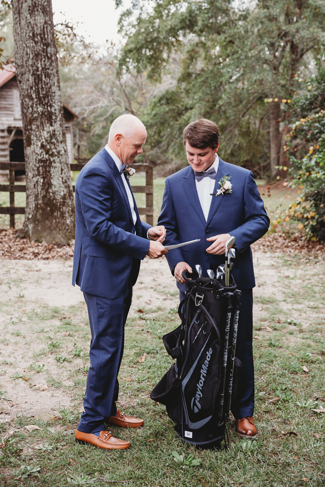 Tender moment as the groom and father handle golf clubs in a black TaylorMade bag under tall oak trees at Wavering Place in Eastover SC.