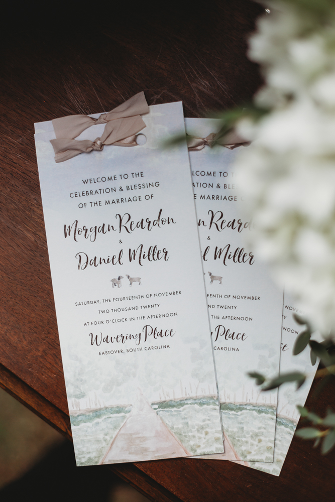 Close-up of watercolor ceremony programs with taupe ribbon bows on a wooden table for a wedding at Wavering Place in Eastover SC.