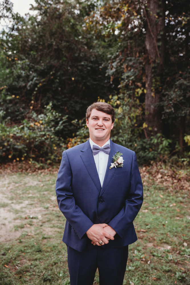 The groom smiles in a navy suit with bowtie and boutonnière, standing on grass with leafy trees behind at Wavering Place in Eastover SC.