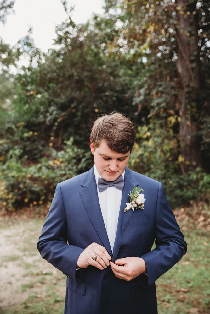 The groom buttons his navy suit jacket while looking down, showcasing the boutonnière and soft fall lighting at Wavering Place in Eastover SC.