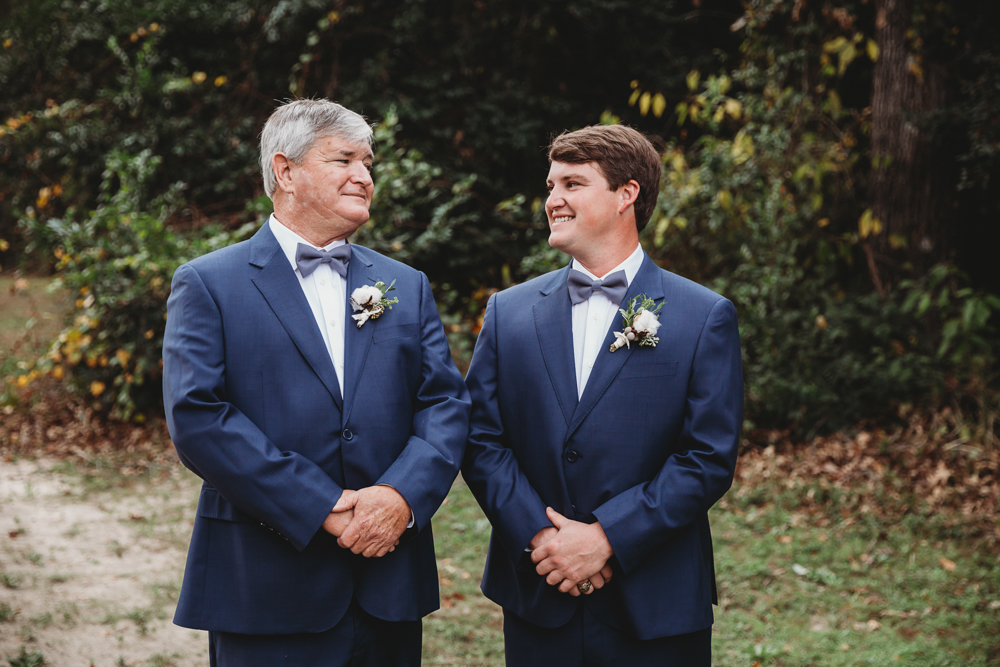The groom and his father share a proud moment in matching blue suits and bowties against the wooded background at Wavering Place in Eastover SC.