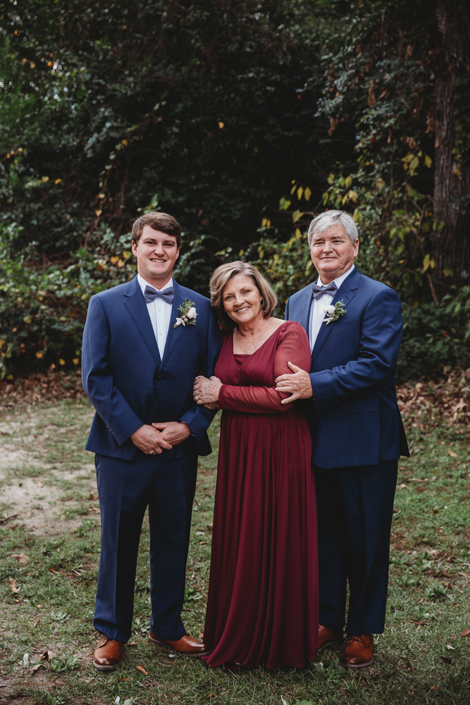 The groom poses with his mom in a wine-colored dress and dad in a navy suit on grass with natural fall foliage behind at Wavering Place in Eastover SC.