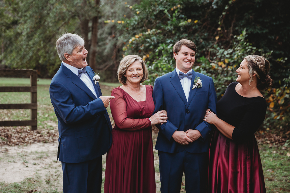 The groom laughs with his parents and sister in coordinated navy and wine tones during a playful candid at Wavering Place in Eastover SC.