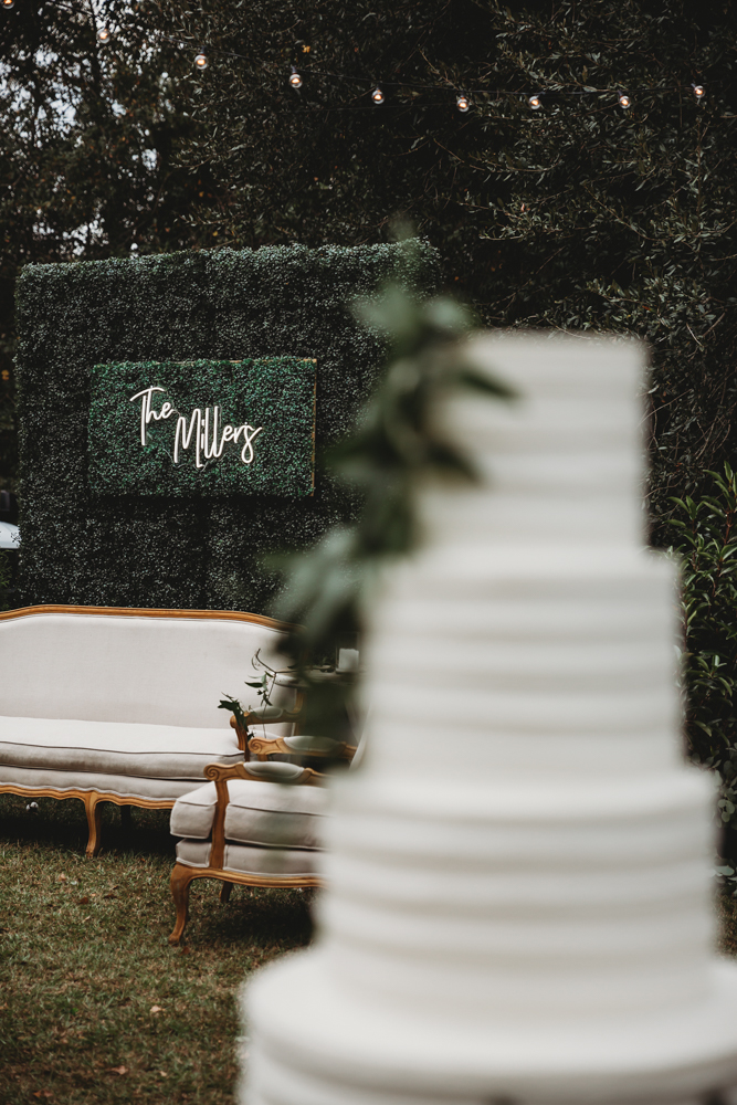 Boxwood greenery wall with 'The Millers' neon sign and vintage white lounge seating next to the wedding cake
