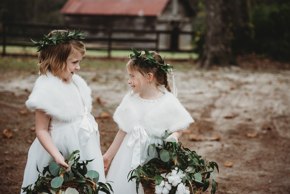 Two flower girls in white dresses with faux fur wraps and greenery crowns hold cotton-filled baskets