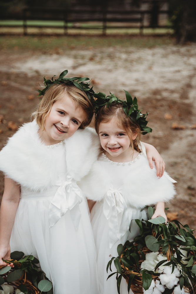 Adorable flower girls pose arm-in-arm in matching white dresses and floral crowns, smiling sweetly