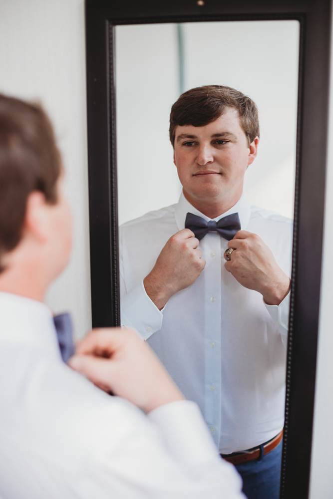 Groom adjusts his bow tie in the mirror during wedding day preparations
