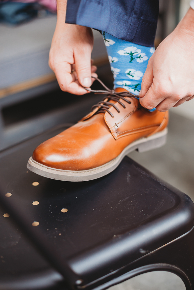 Groom tying laces on his tan leather dress shoes, wearing playful blue socks with cotton motifs