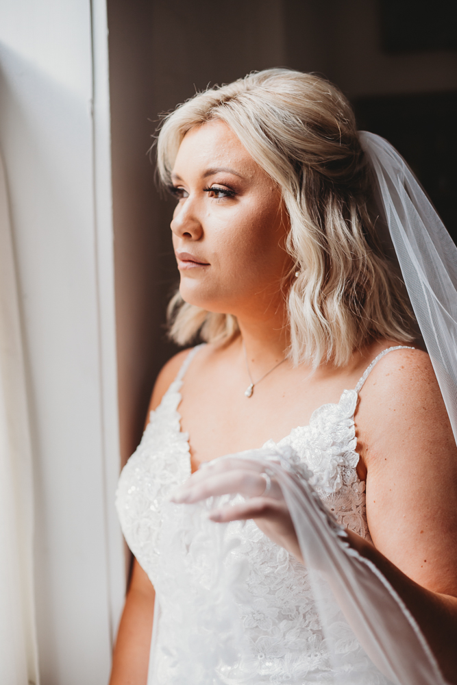 The bride stands in soft window light, gently holding her veil with a peaceful expression before walking down the aisle.
