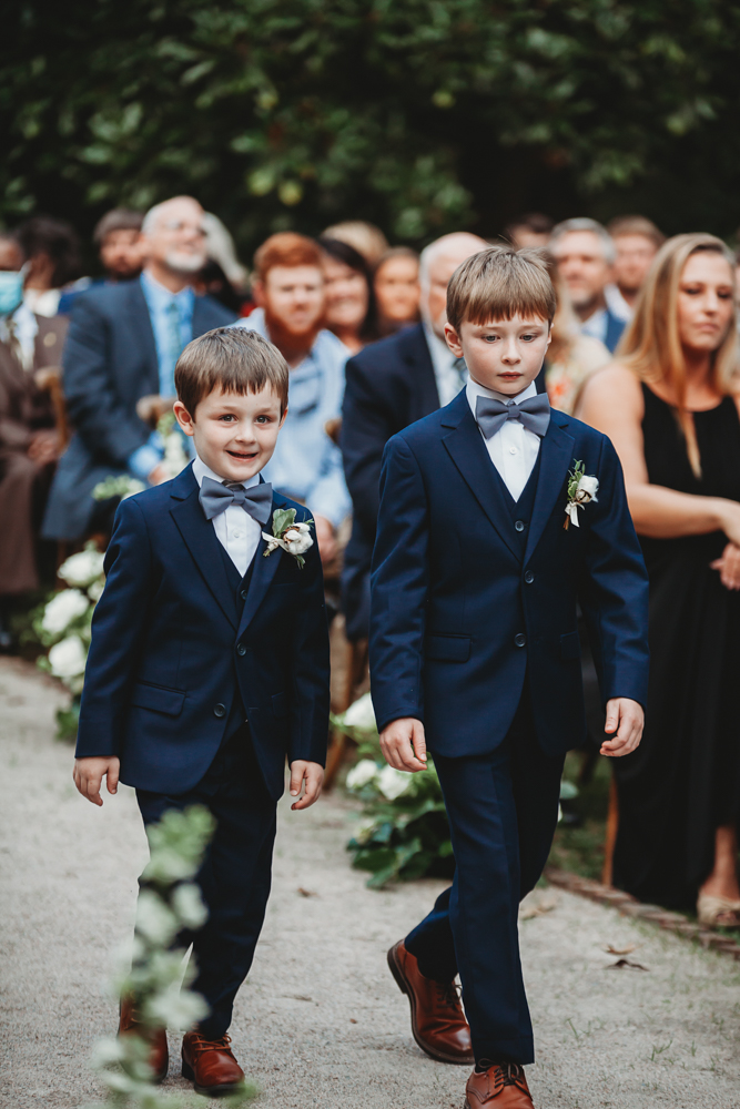 Two ring bearers in navy suits and brown shoes walk together down the flower-lined aisle at Wavering Place.