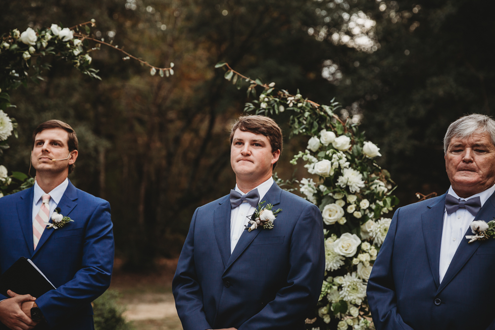 Groom Daniel stands at the altar under white floral arrangements, awaiting his bride with an emotional expression.