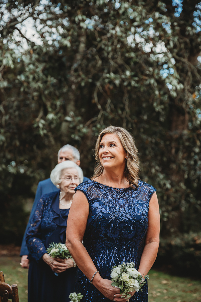The mother of the groom walks with his grandparents down the aisle, all smiling warmly during the wedding processional.