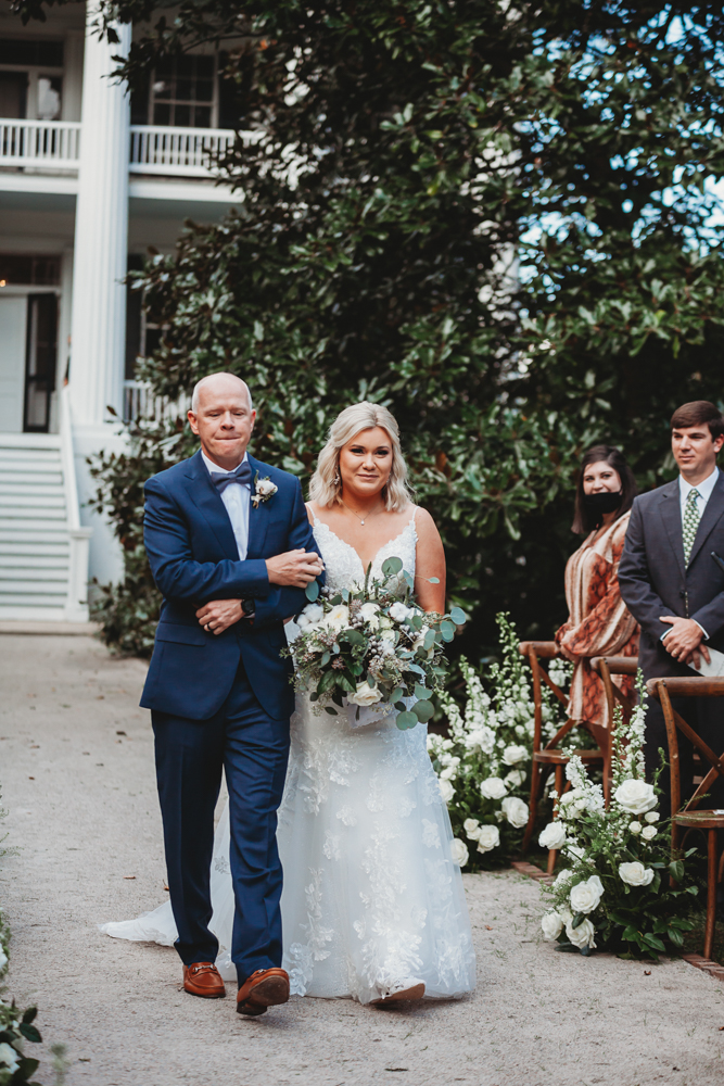 Bride Morgan and her father walk arm-in-arm down a flower-lined aisle as guests watch during the wedding ceremony at Wavering Place.
