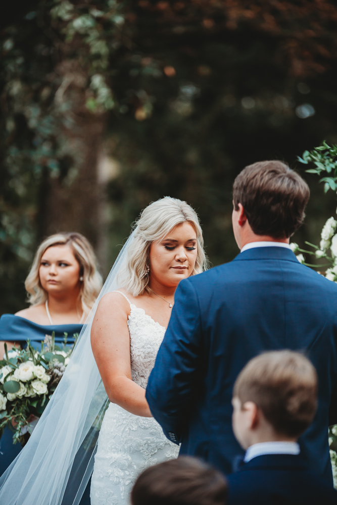 Close-up of Morgan during vows with veil draped over lace gown, Daniel in navy blue suit, and soft florals in hand at Reflection Images by Tracy Rowell in Florence SC