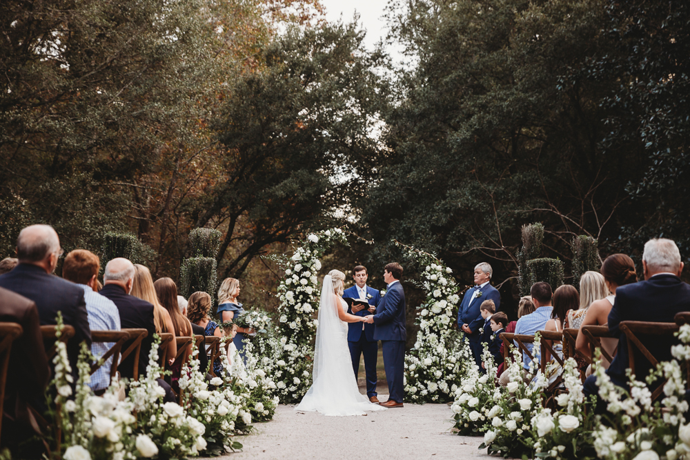 Full ceremony view at Wavering Place with aisle florals, live oak canopy, and guests seated in rows as Morgan and Daniel exchange vows at Reflection Images by Tracy Rowell in Florence SC