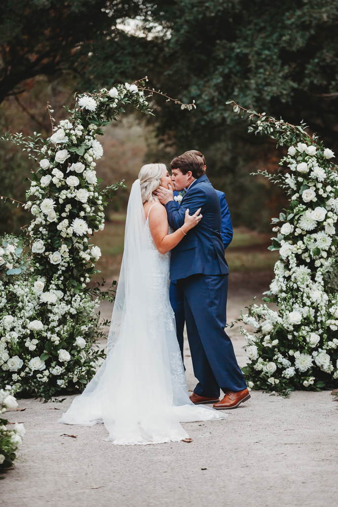Morgan and Daniel's first kiss under lush white floral arch surrounded by greenery at their Wavering Place ceremony, captured by Reflection Images by Tracy Rowell in Florence SC