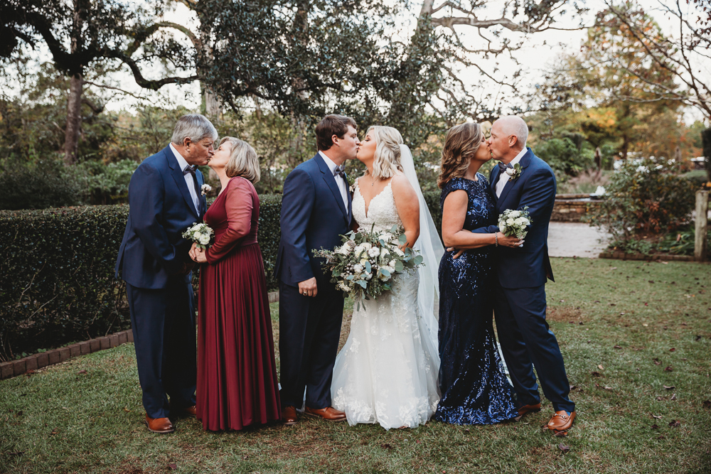 Morgan and Daniel kissing between both sets of parents sharing kisses in formal wear on the lawn at Wavering Place in Eastover, South Carolina