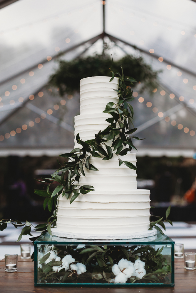 White tiered wedding cake adorned with greenery and displayed over cotton-filled glass box under tented reception lights at Wavering Place, captured by Reflection Images by Tracy Rowell in Florence SC