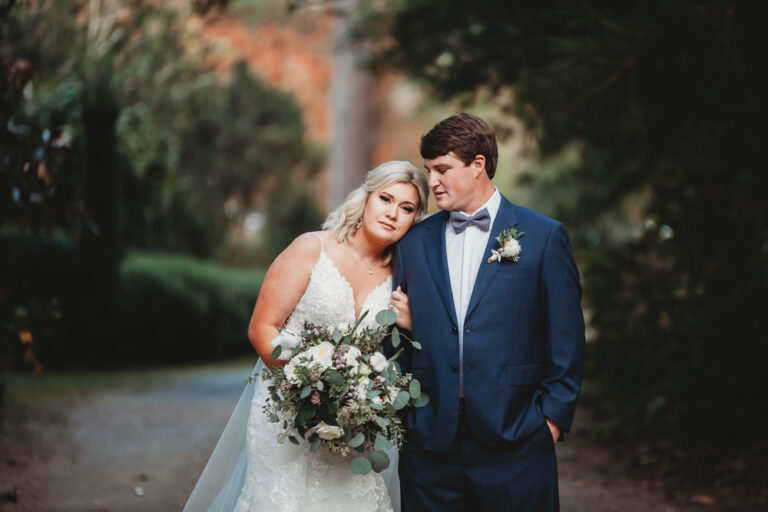 Morgan resting her head on Daniel's shoulder while holding her bouquet, framed by lush trees in soft natural light at Wavering Place