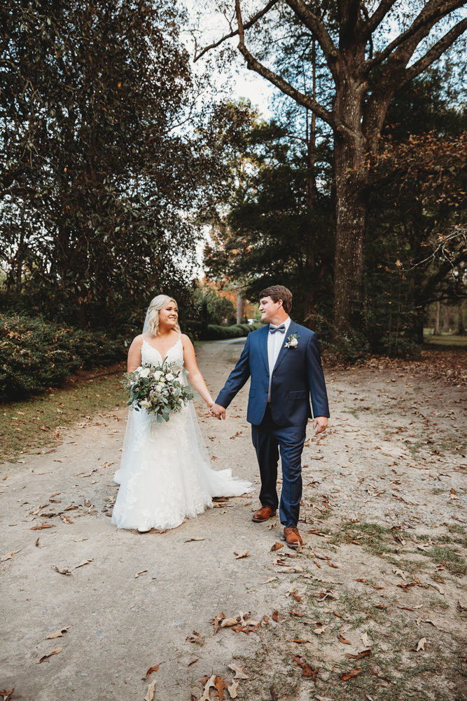 Bride and groom walking hand in hand down a leaf-covered path, smiling at each other in their formal wedding attire at Wavering Place