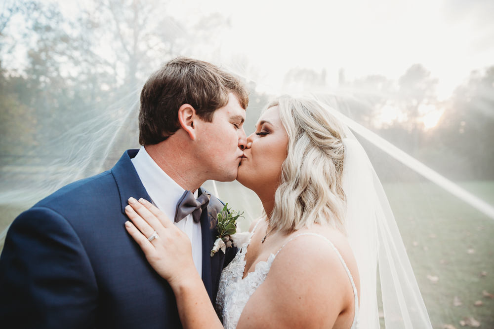 Morgan and Daniel share a kiss beneath her veil with golden sun peeking through soft trees at Wavering Place in Eastover, SC