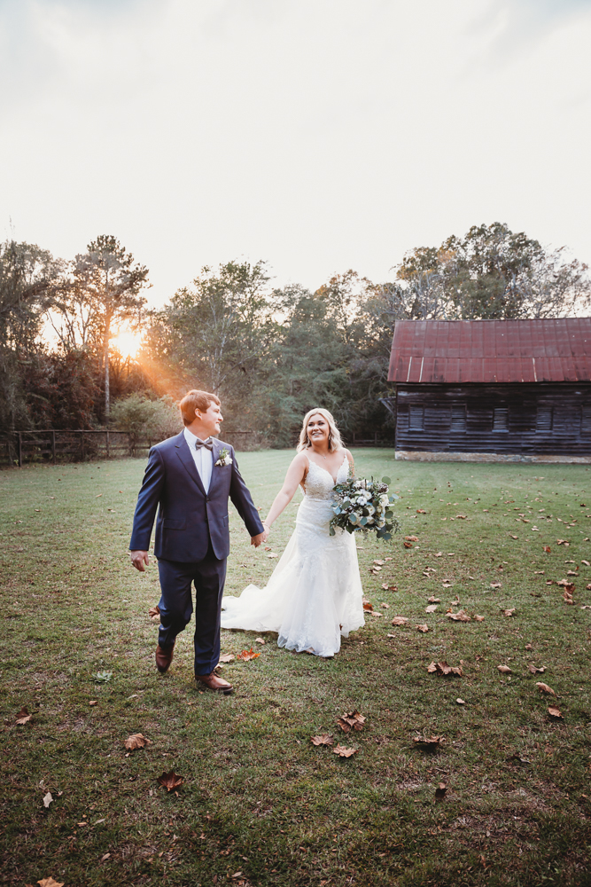 Bride and groom walk across a fall field toward an old barn with sunlight glowing in the background at Wavering Place