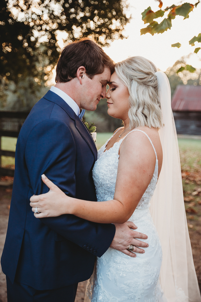 Morgan and Daniel standing forehead to forehead beneath tall trees with autumn light shining behind them at Wavering Place